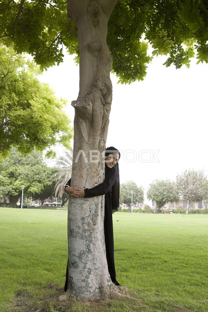 صورة الجسم بالطول الكامل, قضاء أوقات ممتعة في إحدى الحدائق العامة, شابة عربية خليجية إماراتية محجبة تقوم بعناق جذع شجره, اللعب والترفيه في االهواء الطلق, التنزه في الحدائق الخضراء, الاستمتاع في التسلية والترفيه في المنتزهات, اللعب في أوقات الفراغ