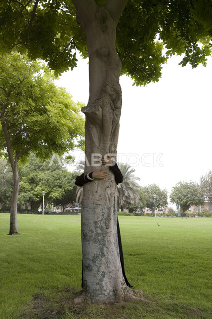 اللعب في أوقات الفراغ, قضاء أوقات ممتعة في إحدى الحدائق العامة, شابة عربية خليجية إماراتية محجبة تقوم بعناق جذع شجره, اللعب والترفيه في الهواء الطلق, التنزه في الحدائق الخضراء, الاستمتاع في التسلية والترفيه في المنتزهات, صورة الجسم بالطول الكامل