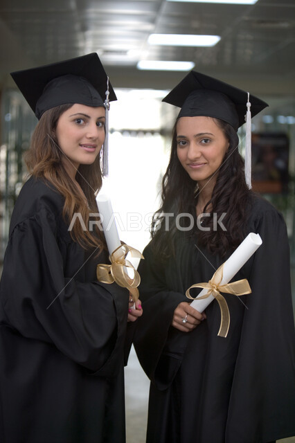 الفرح بتحقيق النجاح وأهمية الدراسة الجامعية، التعليم الجامعي الاماراتي، الاحتفال بالتخرج من الجامعة بإيماءات السعادة والفرح، صورة مقربة لخريجتين عربيتين خليجيتين اماراتيتين مبتسمتين يرتديان ملابس التخرج وتمسكان شهادة التفوق