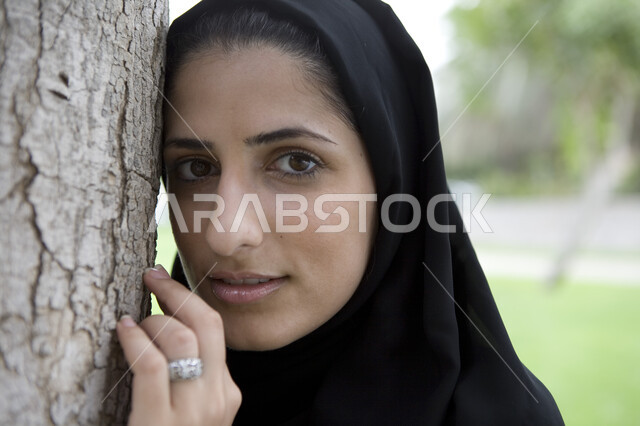 الإستجمام والتنزه في الهواء الطلق, الاستمتاع في التنزه في يوم مشمس, قضاء اوقات ممتعة في إحدى الحدائق العامة, صورة مقربة لشابة عربية خليجية إماراتية ترتدي العباء السوداء محجبة تقوم بالإتكاء على جذع شجرة, عطلة نهاية الاسبوع