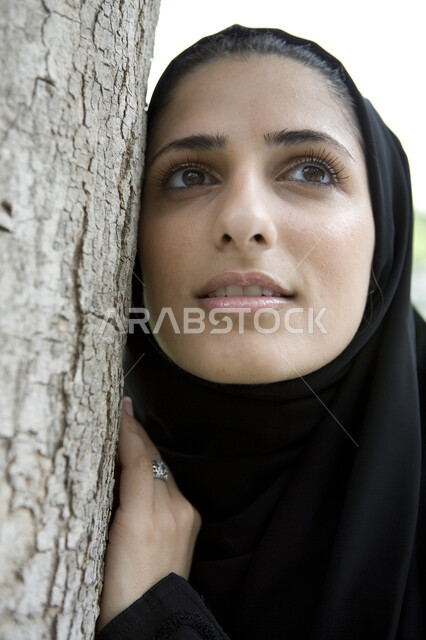 عطلة نهاية الاسبوع, الإستجمام والتنزه في الهواء الطلق, الاستمتاع في التنزه في يوم مشمس, قضاء اوقات ممتعة في إحدى الحدائق العامة, صورة مقربة لشابة عربية خليجية إماراتية ترتدي العباء السوداء محجبة تقوم بالإتكاء على جذع شجرة