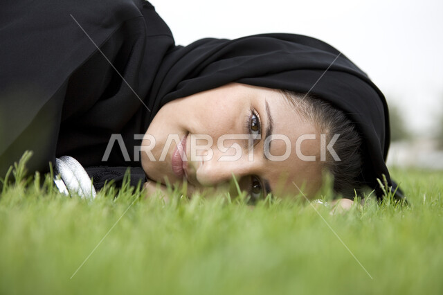 الاستمتاع في التنزه في يوم مشمس, قضاء اوقات ممتعة في إحدى الحدائق العامة, الإستجمام والتنزه في الهواء الطلق, صورة مقربة لشابة عربية خليجية إماراتية ترتدي العباء السوداء محجبة تقوم بالإستلقاء على العشب الاخضر, عطلة نهاية الاسبوع