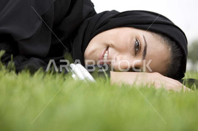 قضاء اوقات ممتعة في إحدى الحدائق العامة, صورة مقربة لشابة عربية خليجية إماراتية ترتدي العباء السوداء محجبة تقوم بالإستلقاء على العشب الاخضر, عطلة نهاية الاسبوع, الإستجمام والتنزه في الهواء الطلق, الاستمتاع في التنزه في يوم مشمس