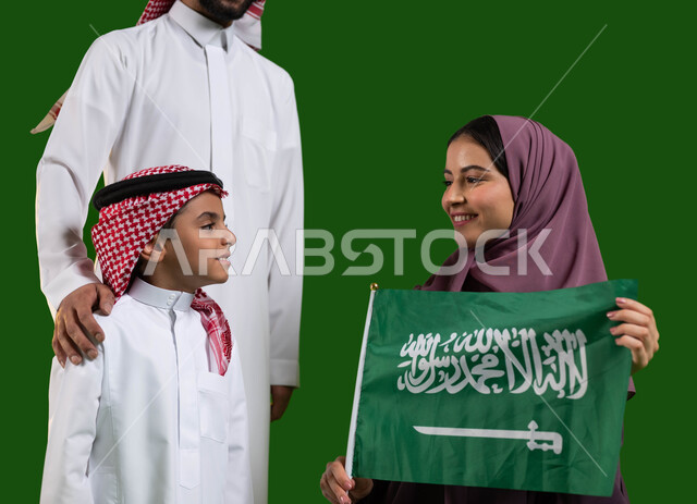 ترسيخ جذور الانتماء للمملكة، الاحتفال مع الأبناء باليوم الوطني 8 ربيع الاول، بورتريه مقرب لعائلة عربية خليجية سعودية سعيدة، ام تمسك علم السعودية بايماءات الفخر والاعتزاز بالوطن، يوم العلم السعودي 11 مارس، العيد الوطني السعودي، كروما، خلفية خضراء