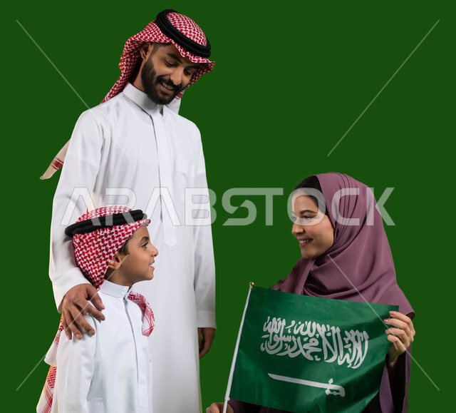 الاحتفال مع الأبناء باليوم الوطني السعودي، بورتريه لعائلة عربية خليجية سعودية سعيدة، الولاء للوطن والمشاركة في المناسبات الوطنية، ام تمسك علم السعودية بايماءات الفخر والاعتزاز، يوم العلم السعودي 11 مارس، العيد الوطني 23 سبتمبر، كروما، خلفية خضراء