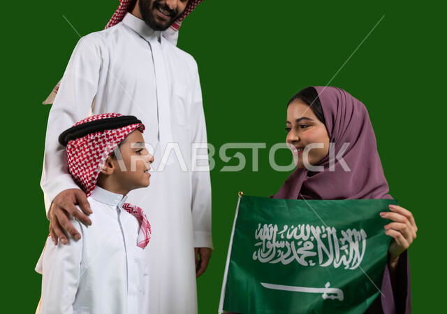 Teaching children to appreciate and love the homeland, the concept of devotion and loyalty to the Kingdom, a portrait of a happy Saudi Gulf Arab family, a mother holding the Saudi flag with gestures of pride and pride, Saudi Flag Day March 11, Saudi National Day, National Day 8 Rabi’ al-Awwal, chroma, green background