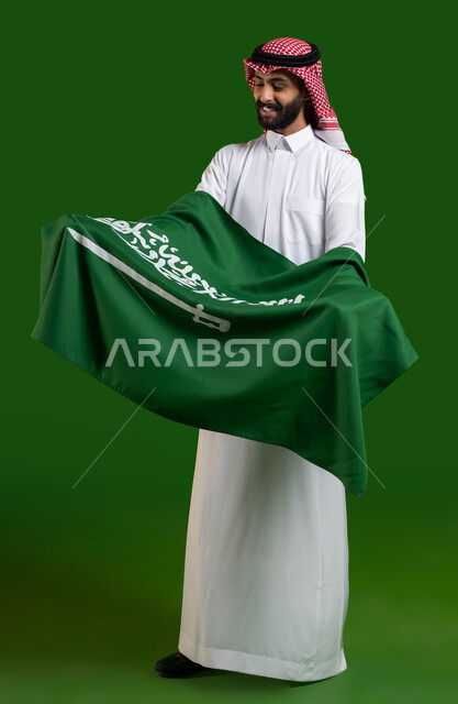 تعظيم كلمة التوحيد على العلم السعودي، بورتريه لمواطن عربي خليجي سعودي يرتدي الشماغ والثوب التقليدي ويحمل علم السعودية بين ذراعيه باحترام وتقدير، اليوم الوطني السعودي، يوم العلم 11 مارس، العيد الوطني 8 ربيع الأول، صورة الجسم بالطول الكامل، كروما، خلفية خضراء