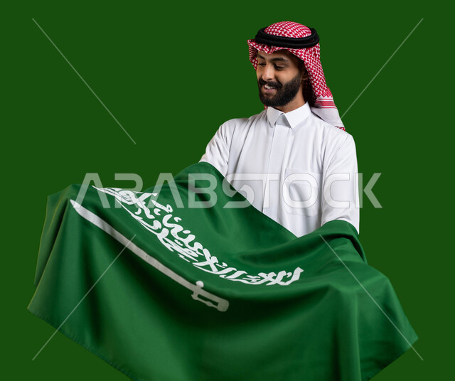 Showing love and loyalty to the state, portrait of a Saudi Gulf Arab man wearing traditional clothing and holding the Saudi flag in his arms with respect and appreciation, glorifying the word monotheism on the Saudi flag, National Day for the Unification of the Kingdom, Flag Day March 11, National Day September 23, chroma, green background