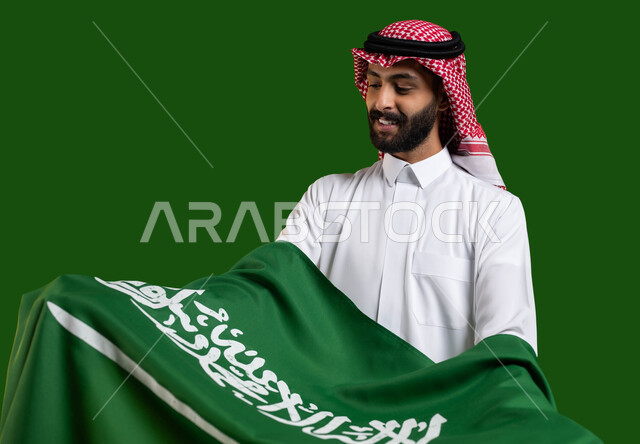 حب الوطن والانتماء إليه، بورتريه لمواطن عربي خليجي سعودي يرتدي الزي التقليدي ويحمل علم السعودية بين ذراعيه باحترام وتقدير، تعظيم كلمة التوحيد على العلم السعودي، اليوم الوطني لتوحيد المملكة، يوم العلم 11 مارس، الاحتفال بالعيد الوطني 23 سبتمبر، كروما، خلفية خضراء
