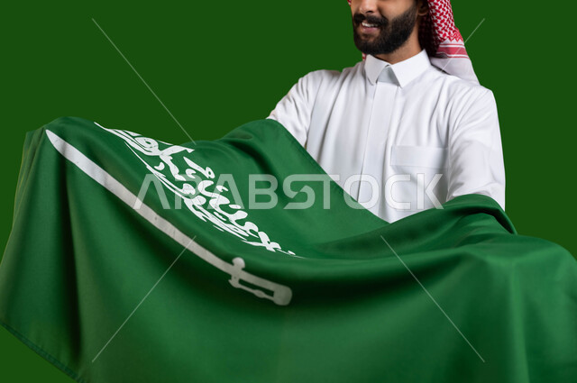 إظهار مشاعر الحب والولاء للمملكة، بورتريه مقرب لشاب عربي خليجي سعودي يرتدي الزي التقليدي ويحمل علم السعودية بين ذراعيه باحترام وتقدير، تعظيم كلمة التوحيد على العلم السعودي، اليوم الوطني السعودي، يوم العلم 11 مارس، العيد الوطني 8 ربيع الأول، كروما، خلفية خضراء