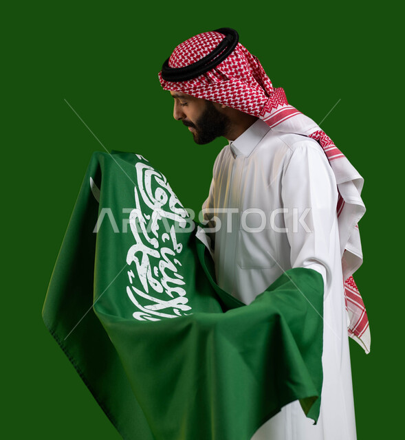 ترسيخ جذور الانتماء الوطني، بورتريه لشاب عربي خليجي سعودي يرتدي الشماغ والثوب التقليدي ويحمل علم السعودية بين ذراعيه باحترام وتقدير، النظر الى العلم عن قرب، اليوم الوطني السعودي، يوم العلم 11 مارس، العيد الوطني 8 ربيع الأول، كروما، خلفية خضراء