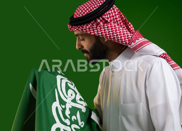 ايماءات الفخر بالهويه الوطنية، بورتريه مقرب لرجل عربي خليجي سعودي يرتدي الشماغ والثوب التقليدي ويحمل علم السعودية بين ذراعيه باحترام وتقدير، النظر الى العلم عن قرب، اليوم الوطني السعودي، يوم العلم 11 مارس، العيد الوطني 8 ربيع الأول، كروما، خلفية خضراء