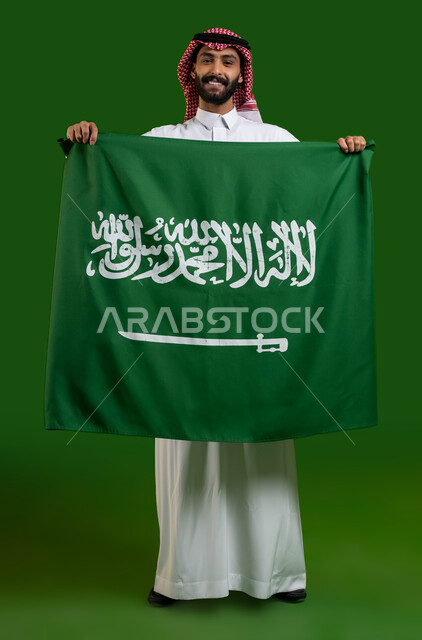 ايماءات السعاده والفخر بالانتماء للهوية الوطنية السعودية، بورتريه لرجل عربي خليجي سعودي مبتسم يرتدي الشماغ والثوب التقليدي ويمسك علم المملكة، اليوم الوطني  23 سبتمبر، يوم العلم 11 مارس، العيد الوطني السعودي، صورة الجسم بالطول الكامل، كروما، خلفية خضراء