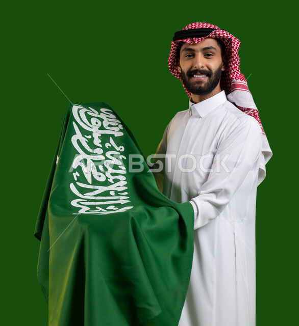 مشاعر الانتماء والحب للبلاد، بورتريه لرجل عربي خليجي سعودي مبتسم يرتدي الزي التقليدي ويحمل علم السعودية بين ذراعيه باحترام وتقدير، تعظيم كلمة التوحيد على العلم السعودي، اليوم الوطني السعودي، يوم العلم 11 مارس، العيد الوطني 23 سبتمبر، كروما، خلفية خضراء