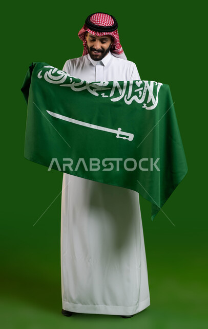 الفخر والتباهي بالإنتماء للمملكة، بورتريه من الخلف لرجل عربي خليجي سعودي يرتدي الزي التقليدي ويحمل علم السعودية وينظر اليه بفخر وتباهي، ذكرى اليوم الوطني 23 سبتمبر، العيد الوطني السعودي، يوم العلم السعودي 11 مارس، صورة الجسم بالطول الكامل، كروما، خلفية خضراء