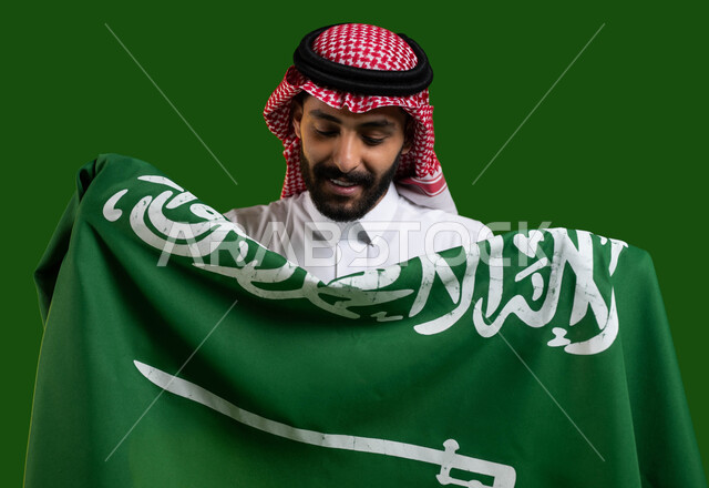 الاحتفال باليوم الوطني لتوحيد السعودية، تعميق الانتماء والولاء للوطن، بورتريه مقرب لشاب عربي خليجي سعودي يرتدي الشماغ والثوب التقليدي ويحمل علم السعوديه وينظر اليه بفخر وتباهي، العيد الوطني السعودي 23 سبتمبر، يوم العلم السعودي 11 مارس، كروما، خلفية خضراء