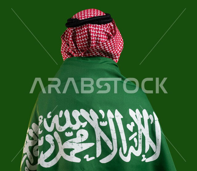 The memory of the National Day for the unification of the Kingdom, expressing feelings of pride in the homeland, a portrait from behind of a Saudi Arabian Gulf youth wearing the Saudi flag on his back, celebrating Flag Day March 11, the Saudi National Day 8 Rabi` al-Awwal, participation in national events and holidays, chrome, green background