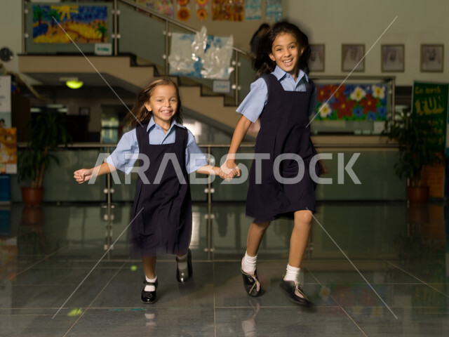وقت اللعب والترفيه أثناء الفسحة المدرسية، التعليم في الإمارات العربية المتحدة، الاستعداد لبداية فصل دراسي جديد، طالبتان عربيتان خليجيتان إماراتيتان ترتديان الزي المدرسي تستمتعان بالركض في بهو المدرسة، إيماءات تدل على السعادة والمرح.