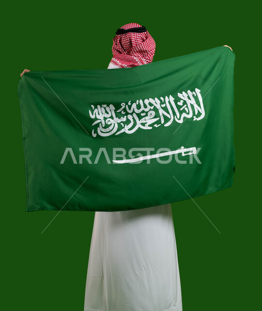 الفخر والاعتزاز بالهوية الوطنية، بورتريه من الخلف لرجل عربي خليجي سعودي يرتدي الزي التقليدي ويحمل علم السعودية، تعميق الانتماء للوطن بالاحتفال باليوم الوطني لتوحيد السعودية، العيد الوطني السعودي 8 ربيع الأول، يوم العلم السعودي 11 مارس، كروما، خلفية خضراء