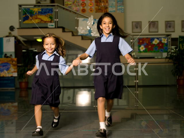 وقت اللعب والترفيه أثناء الفسحة المدرسية، التعليم في الإمارات العربية المتحدة، الاستعداد لبداية فصل دراسي جديد، طالبتان عربيتان خليجيتان إماراتيتان ترتديان الزي المدرسي تستمتعان بالركض في بهو المدرسة، إيماءات تدل على السعادة والمرح.