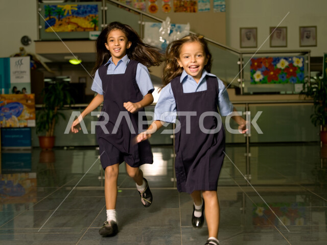 وقت اللعب والترفيه أثناء الفسحة المدرسية، التعليم في الإمارات العربية المتحدة، الاستعداد لبداية فصل دراسي جديد، طالبتان عربيتان خليجيتان إماراتيتان ترتديان الزي المدرسي تستمتعان بالركض في بهو المدرسة، إيماءات تدل على السعادة والمرح.