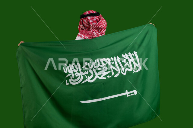 إظهار مشاعر الحب والولاء للمملكة، بورتريه من الخلف لمواطن عربي خليجي سعودي يرتدي الزي التقليدي ويحمل علم السعودية، الاحتفال بذكرى اليوم الوطني السعودي، العيد الوطني 23 مارس، يوم العلم السعودي 11 مارس، التضحية والوفاء للمملكة، كروما، خلفية خضراء