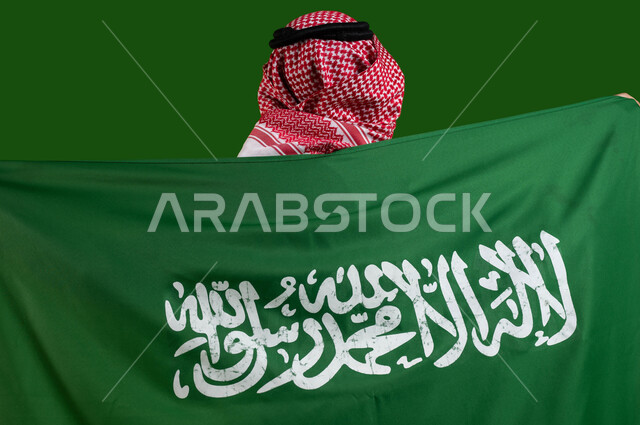 مفهوم الولاء للوطن والمشاركة بالمناسبات و الأعياد الوطنية، بورتريه من الخلف لشاب عربي خليجي سعودي يرتدي الزي التقليدي ويحمل علم السعودية، ذكرى اليوم الوطني لتوحيد المملكة، العيد الوطني السعودي 8 ربيع الأول، يوم العلم 11 مارس، كروما، خلفية خضراء