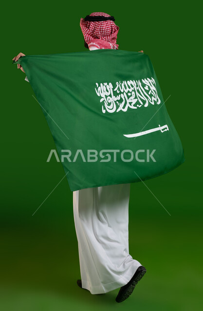Pride and boasting of belonging to the Kingdom, a portrait from behind of a Saudi Arabian Gulf man wearing traditional dress and carrying the Saudi flag, the anniversary of the National Day for the Unification of Saudi Arabia, the Saudi National Day March 23, the Saudi Flag Day March 11, full-length body image, chroma, green background