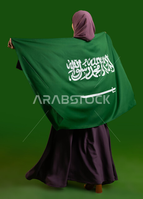 يوم العلم السعودي 11 مارس، تعميق الانتماء والولاء للوطن، بورتريه من الخلف لامرأة عربية خليجية سعودية محجبة تضع علم السعودية على ظهرها، الإحتفال بالعيد الوطني 8 ربيع الأول، ذكرى اليوم الوطني السعودي، صورة الجسم بالطول الكامل، كروما، خلفية خضراء