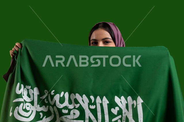 الإحتفال بيوم العلم 11 مارس، بورتريه لسيدة عربية سعودية خليجية محجبة تقوم بتغطية وجهها بعلم المملكة العربية السعودية وتنظر الى الكاميرا، ذكرى اليوم الوطني السعودي 8 ربيع الأول، العيد الوطني السعودي، التباهي والفخر بالهوية الوطنية، كروما، خلفية خضراء