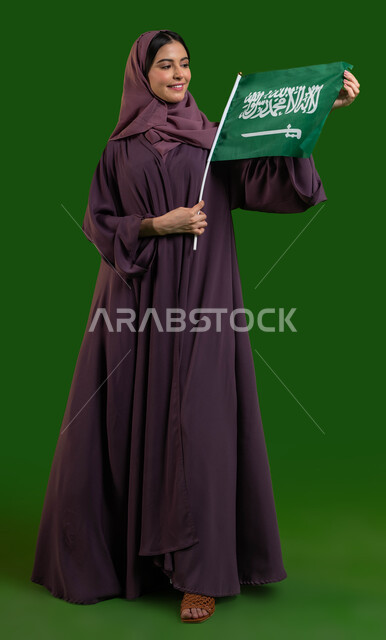 بورتريه لسيدة عربية خليجية سعودية محجبة تسمك العلم السعودي وتنظر إليه بايماءات الفخر والاعتزاز بالوطن، العيد الوطني السعودي، اليوم الوطني 8 ربيع الأول، يوم العلم 11 مارس، مفهوم الولاء للوطن والمشاركة بالأعياد الوطنية، صورة الجسم بالطول الكامل، كروما، خلفية خضراء