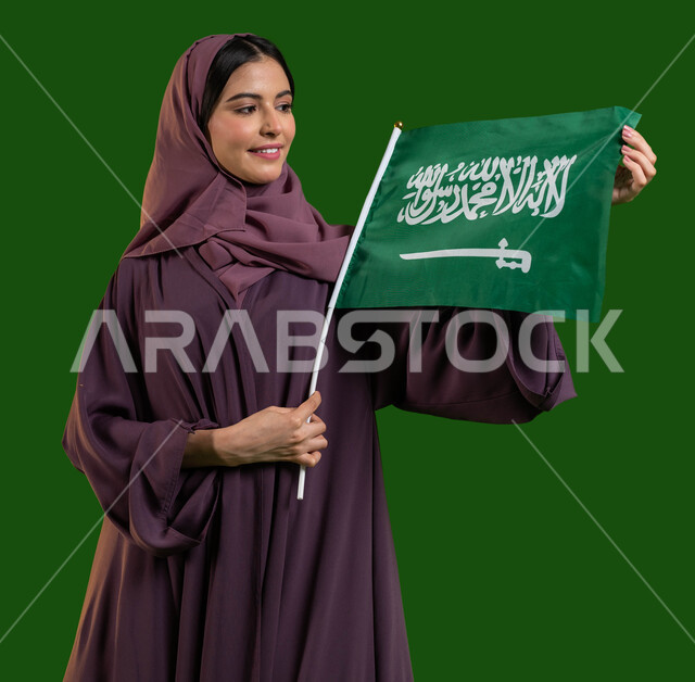 الإنتماء للوطن وحب البلاد، بورتريه لامرأة عربية خليجية سعودية محجبة تسمك علم السعودية وتنظر إليه بايماءات الفخر والاعتزاز بالوطن، اليوم الوطني السعودي، اليوم الوطني 23 سبتمبر، يوم العلم السعودي 11 مارس، الاحتفال بالأعياد والمناسبات الوطنية، كروما، خلفية خضراء