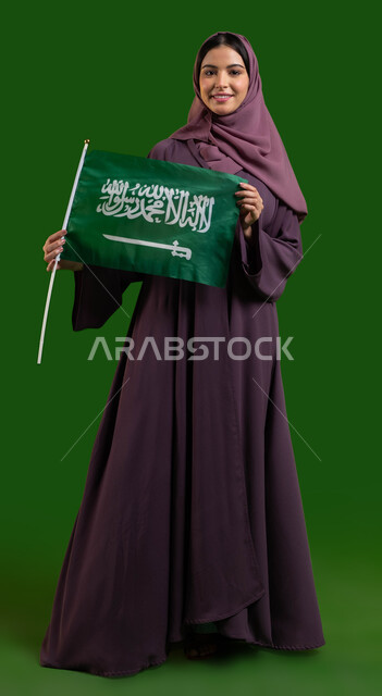 اليوم الوطني السعودي، المشاركة الأعياد الوطنية، بورتريه لامرأة عربية خليجية سعودية محجبة مبتسمة ترتدي الحجاب وترفع علم السعودية بايماءات الفخر والاعتزاز بالوطن، يوم العلم السعودي 11 مارس، العيد الوطني 23 سبتمبر، صورة الجسم بالطول الكامل، كروما، خلفية خضراء