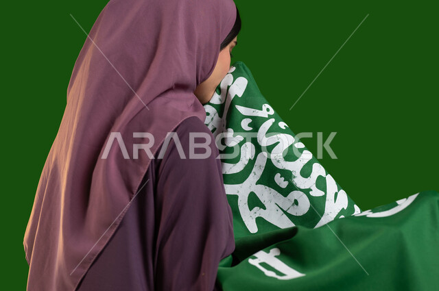 الاعتزاز والفخر بالهوية الوطنية، بورتريه مقرب من الجانب لمواطنة عربية خليجية سعودية محجبة تقوم بتقبيل علم المملكة، يوم العلم السعودي 11 مارس، الاحتفال باليوم الوطني السعودي 23 سبتمبر، العيد الوطني لتوحيد السعودية، كروما، خلفية خضراء