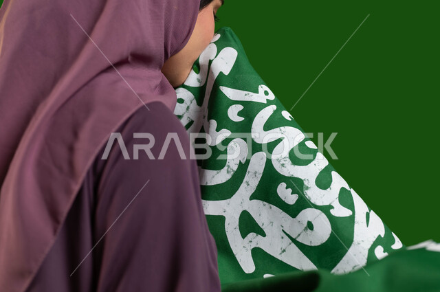 مفهوم حب الوطن والاعتزاز به، بورتريه مقرب من الجانب لسيدة عربية خليجية سعودية محجبة تقوم بتقبيل علم المملكة، يوم العلم 11 مارس، اليوم الوطني السعودي، الاحتفال بالعيد الوطني السعودي 8 ربيع الأول، كروما، خلفية خضراء