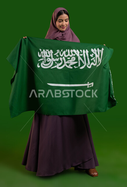 التعبير عن مشاعر الاعتزاز بالوطن، بورتريه لمواطنة عربية خليجية سعودية محجبة تحمل علم المملكة بفخر وإعتزاز، الاحتفال باليوم الوطني لتوحيد السعودية 8 ربيع الاول، يوم العلم السعودي 11 مارس، العيد الوطني السعودي، صورة الجسم بالطول الكامل، كروما، خلفية خضراء