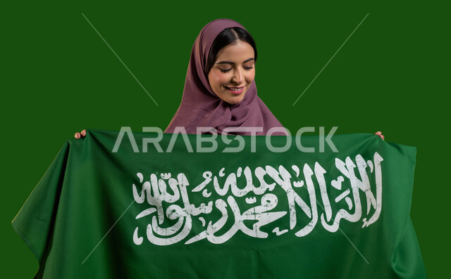 الفخر والاعتزاز بالهوية الوطنية، بورتريه لسيدة عربية خليجية سعودية محجبة تحمل علم المملكة بإيماءات السرور والسعادة، الاحتفال بيوم العلم السعودي 11 مارس، احياء ذكرى اليوم الوطني  لتوحيد السعودية، العيد الوطني السعودي 8 ربيع الأول، كروما، خلفية خضراء