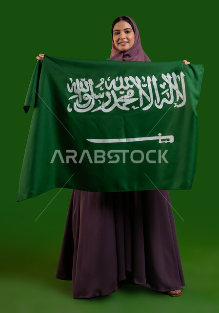 حب الوطن والانتماء إليه، بورتريه لمواطنة عربية خليجية سعودية محجبة تحمل علم المملكة بإيماءات السرور، الاحتفال بيوم العلم 11 مارس، احياء ذكرى اليوم الوطني السعودي، العيد الوطني لتوحيد السعودية 8 ربيع الأول، صورة الجسم بالطول الكامل، كروما، خلفية خضراء
