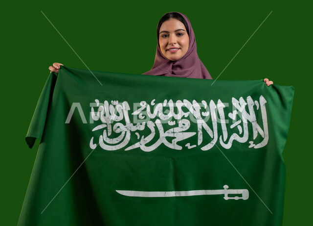 إظهار مشاعر الاعتزاز بالوطن، بورتريه لسيدة عربية خليجية سعودية محجبة تحمل علم المملكة بإيماءات السرور والسعادة، الاحتفال بيوم العلم السعودي 11 مارس، احياء ذكرى اليوم الوطني السعودي، العيد الوطني لتوحيد السعودية 23 سبتمبر، كروما، خلفية خضراء
