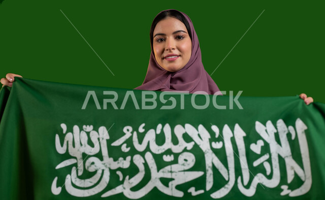 إظهار الحب والولاء للدولة، بورتريه لامرأة عربية خليجية سعودية محجبة  تحمل علم السعودية بإيماءات السرور والسعادة، تعميق الانتماء للوطن بالاحتفال بذكرى اليوم الوطني لتوحيد المملكة 8 ربيع الأول، العيد الوطني السعودي، يوم العلم 11 مارس، كروما، خلفية خضراء