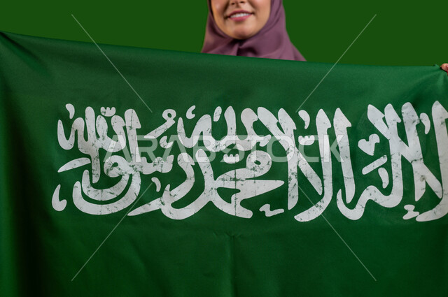 الاعتزاز والفخر بالهوية الوطنية، بورتريه مقرب لمواطنة عربية خليجية سعودية محجبة  تحمل علم السعودية بإيماءات السرور والسعادة، تعميق الانتماء للوطن بالاحتفال بذكرى اليوم الوطني 23 سبتمبر، العيد الوطني السعودي، يوم العلم السعودي 11 مارس، كروما، خلفية خضراء