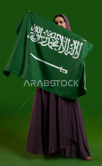 إظهار الحب والولاء للدولة، بورتريه لإمرأة عربية خليجية سعودية محجبة ترتدي العباية وتقوم بتغطية وجهها بعلم السعودية، احياء ذكرى اليوم الوطني السعودي، الاحتفال بالعيد الوطني 23 سبتمبر، يوم العلم السعودي 11 مارس، صورة الجسم بالطول الكامل، كروما، خلفية خضراء
