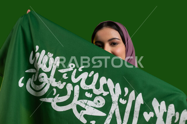 الفخر والتباهي بالإنتماء للمملكة، بورتريه لسيدة عربية خليجية سعودية محجبة تقوم بتغطية وجهها بعلم السعودية، الإحتفال باليوم الوطني السعودي 23 سبتمبر، ذكرى العيد الوطني لتوحيد السعودية، يوم العلم السعودي 11 مارس، المشاركة بالاعياد الوطنية، كروما، خلفية خضراء