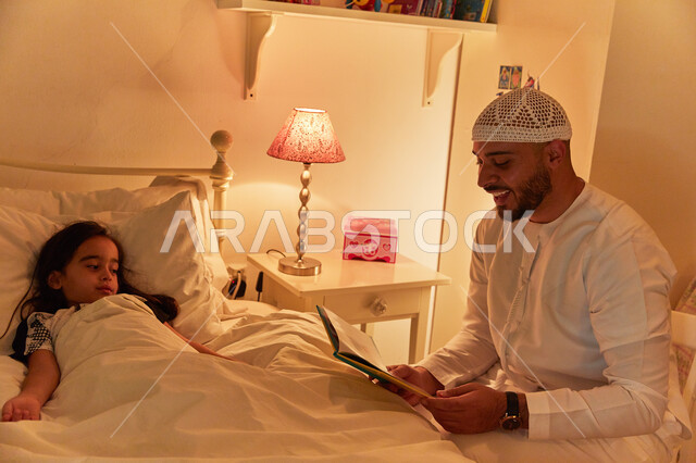 رواية القصص المفيده مع الأبناء قبل النوم، مفهوم التربيه القائمه على الحب والحنان، رجل عربي خليجي إماراتي يرتدي الكندوره والطاقيه الاماراتيه يقوم بقراءة الروايات المسليه لابنته، قضاء وقت ممتع مع الأبنه قبل النوم.