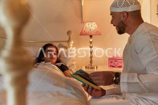 قضاء وقت ممتع مع الأبنه قبل النوم، مفهوم التربيه القائمه على الحب والحنان، رجل عربي خليجي إماراتي يرتدي الكندوره والطاقيه الاماراتيه يقوم بقراءة الروايات المسليه لابنته، رواية القصص المفيده مع الأبناء قبل النوم.