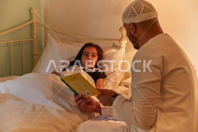التربيه المنزلية القائمه على الحب والحنان، المشاركه بقراءه القصص المفيده مع الأبناء قبل النوم، رجل عربي خليجي إماراتي يرتدي الكندوره والطاقيه الاماراتيه يقرأ الروايات المسليه لابنته، قضاء وقت ممتع مع الأبنه قبل النوم.