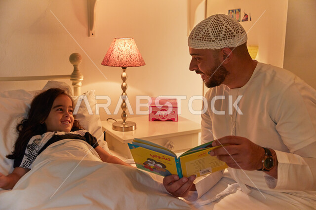 مفهوم الترابط الأسري، مشاركة قراءه الروايات المفيده مع الأبناء قبل النوم، أب عربي خليجي إماراتي يرتدي الكندوره والطاقيه الاماراتيه يقوم بقراءة القصص المسليه لابنته، الشعور بالأمان والحب مع العائله.