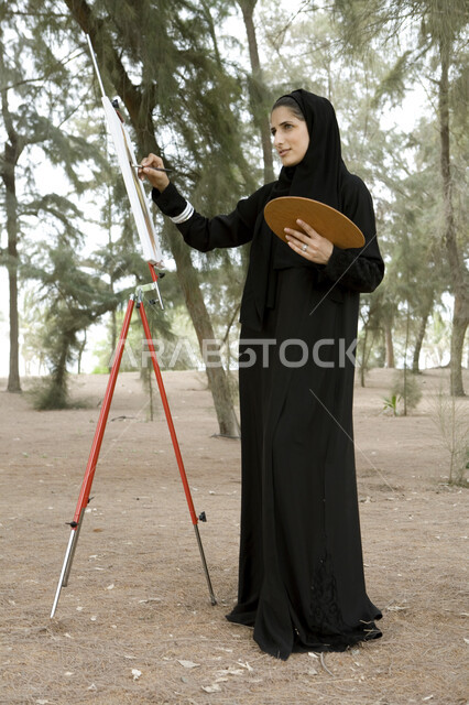 صورة الجسم بالطول الكامل, نشاطات ترفيهية فنية، قضاء اوقات ممتعة في إحدى الحدائق العامة, شابة عربية خليجية إماراتية محجبة تحمل بيدها بالتة ألوان وتقوم بالرسم على اللوحة في المنتزه, ممارسة هواية الرسم والتلوين, الإستجمام والتنزه في الهواء الطلق
