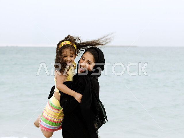 قضاء أوقات عائلية ممتعة بالعطلة الصيفية على شاطئ البحر, صورة مقربة لأم عربية خليجية إماراتية محجبة تحمل إبنتها بحب وحنان، أم تلعب مع إبنتها بإيماءات وجه ويدين مختلفة تدل على السعادة، مفهوم الأمومة والطفولة