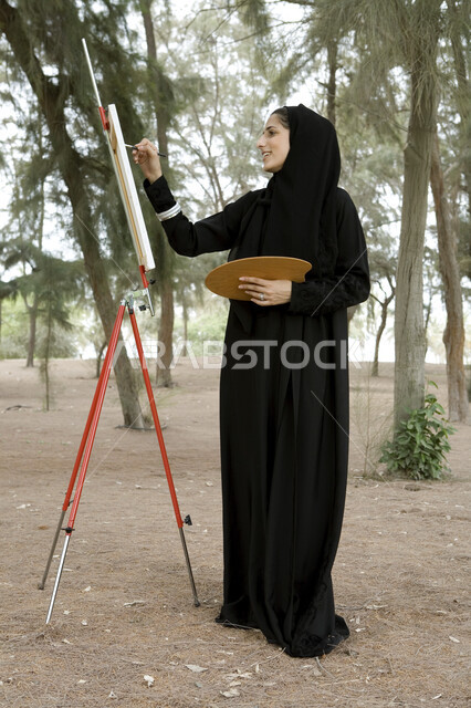 نشاطات ترفيهية فنية، قضاء اوقات ممتعة في إحدى الحدائق العامة, شابة عربية خليجية إماراتية محجبة تحمل بيدها بالتة ألوان وتقوم بالرسم على اللوحة في المنتزه, ممارسة هواية الرسم والتلوين, الإستجمام والتنزه في الهواء الطلق, صورة الجسم بالطول الكامل